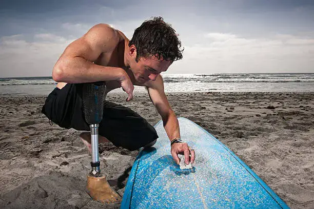 A man with a below-knee prosthesis waxing a surfboard on the beach